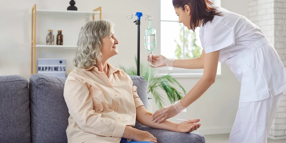 A smiling senior woman sits on a couch as a nurse adjusts her IV drip during IV Therapy in Greenville, SC, in a bright, cozy room.