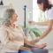 A smiling senior woman sits on a couch as a nurse adjusts her IV drip during IV Therapy in Greenville, SC, in a bright, cozy room.