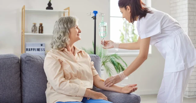 A smiling senior woman sits on a couch as a nurse adjusts her IV drip during IV Therapy in Greenville, SC, in a bright, cozy room.