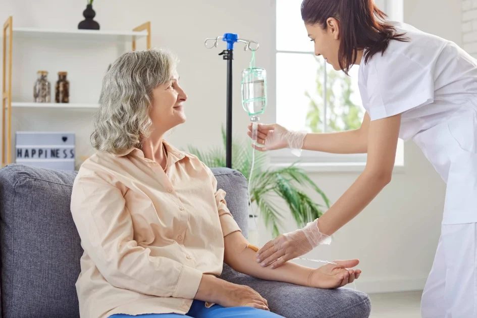 A smiling senior woman sits on a couch as a nurse adjusts her IV drip during IV Therapy in Greenville, SC, in a bright, cozy room.